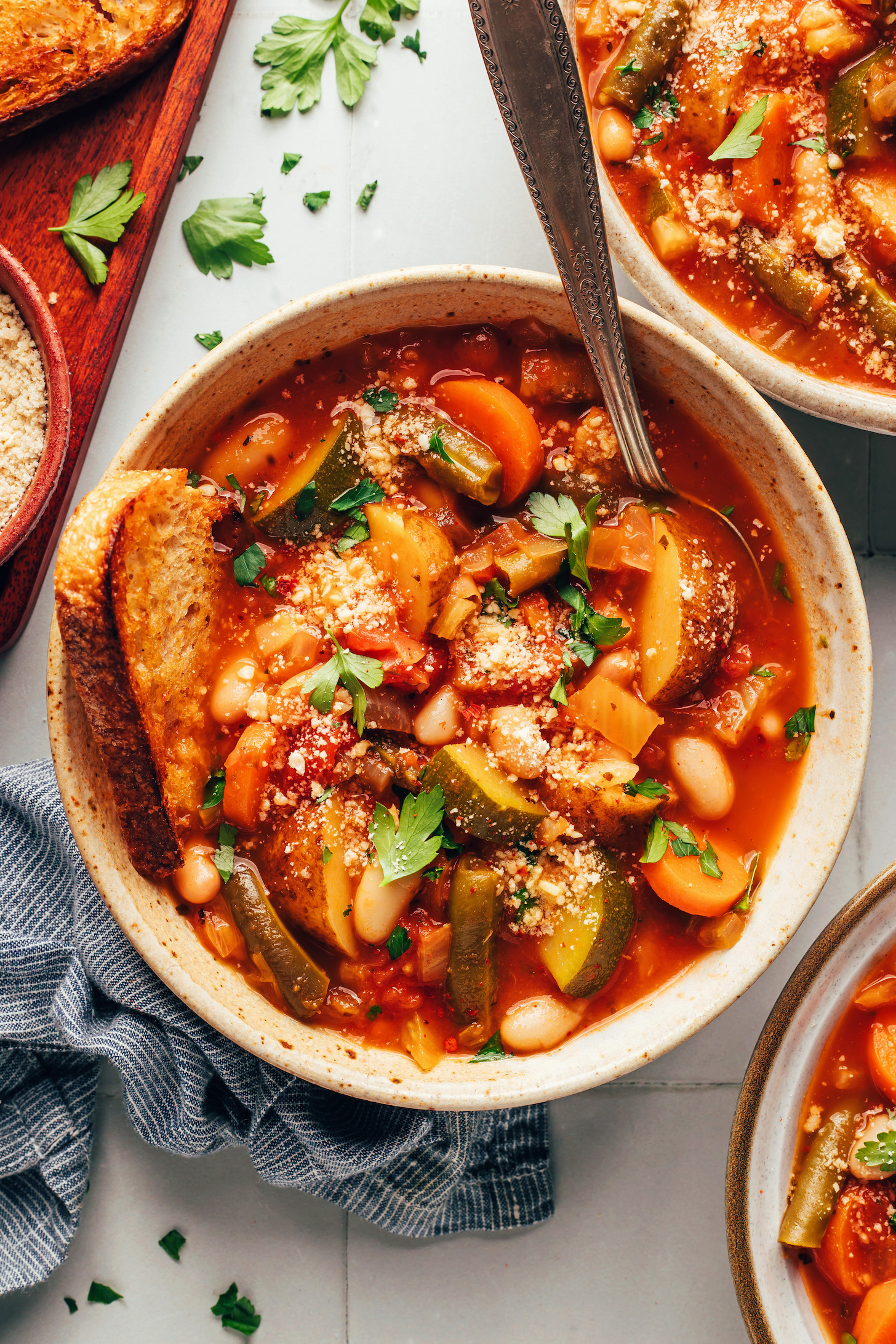 Bowls of veggie white bean minestrone topped with vegan parmesan cheese and parsley and served with toasted bread