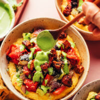 Drizzling pesto sauce onto a bowl of polenta with sheet pan ratatouille and tofu