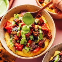 Drizzling pesto sauce onto a bowl of polenta with sheet pan ratatouille and crispy tofu